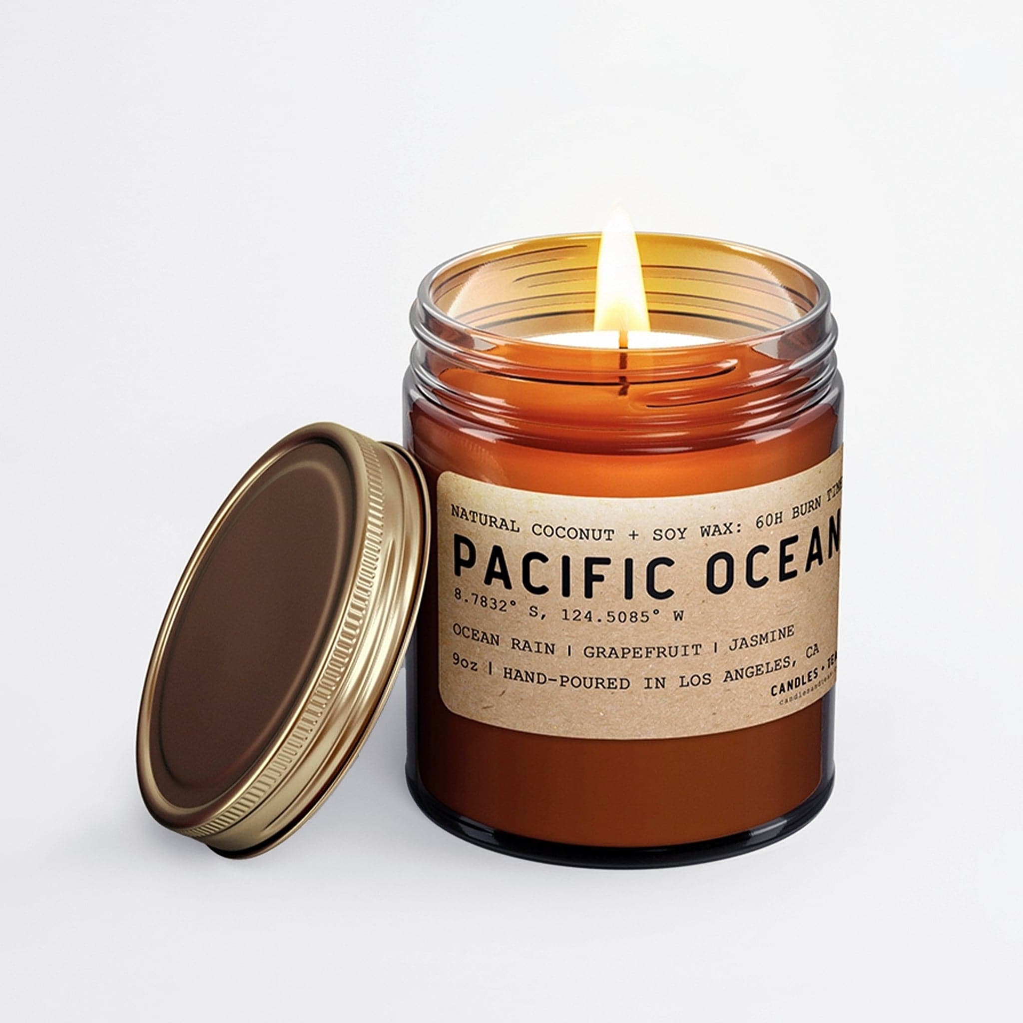Sitting in front of a white background is a dark amber glass jar. Inside the jar is a white candle with a lit wick in the center. On the front is a brown sticker with black text that reads ‘Pacific Ocean.’ There is a gold metal lid leaning against the left side of the jar. 