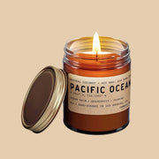 Sitting in front of a white background is a dark amber glass jar. Inside the jar is a white candle with a lit wick in the center. On the front is a brown sticker with black text that reads ‘Pacific Ocean.’ There is a gold metal lid leaning against the left side of the jar.