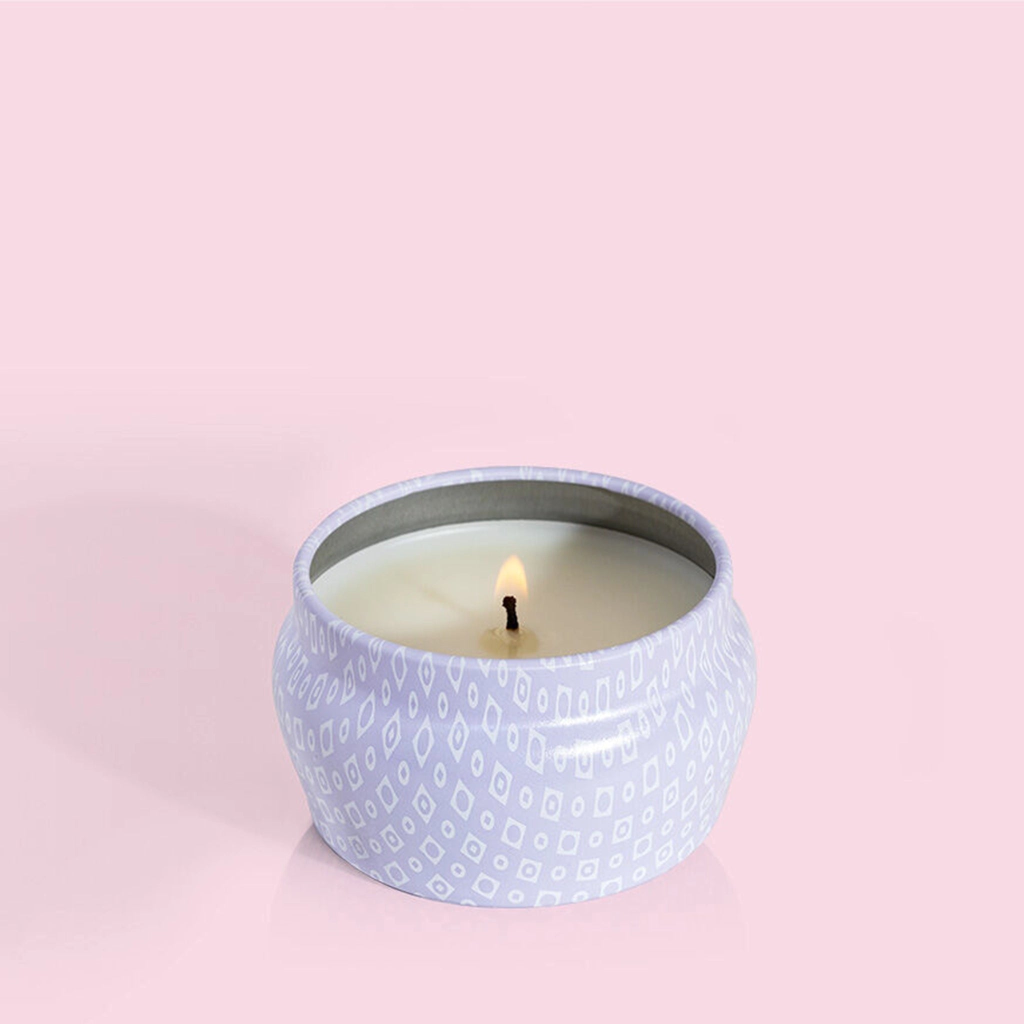 A small lavender tin candle with white wax and a white diamond repeating pattern on the outside of the tin. Comes with a matching purple lid that reads, "Capri Blue Volcano".