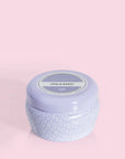 A small lavender tin candle with white wax and a white diamond repeating pattern on the outside of the tin. Comes with a matching purple lid that reads, "Capri Blue Volcano".