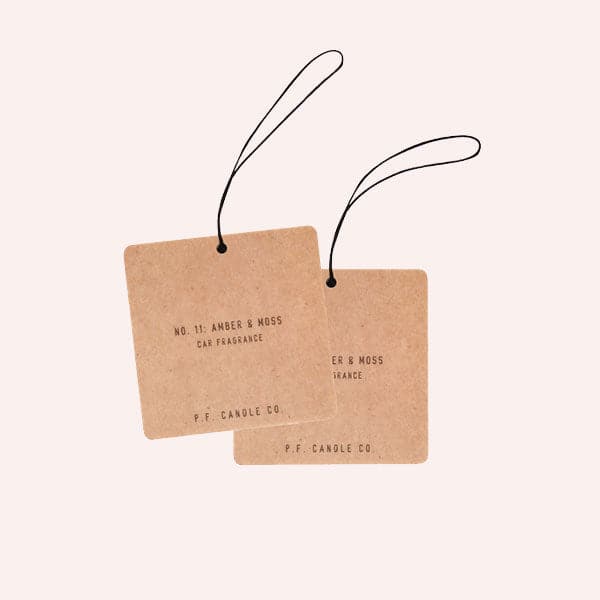 Two square cardboard color air fresheners with an elastic black loop for hanging and small black text that reads, "NO. 11: Amber & Moss Car Fragrance, P.F. Candle Co."