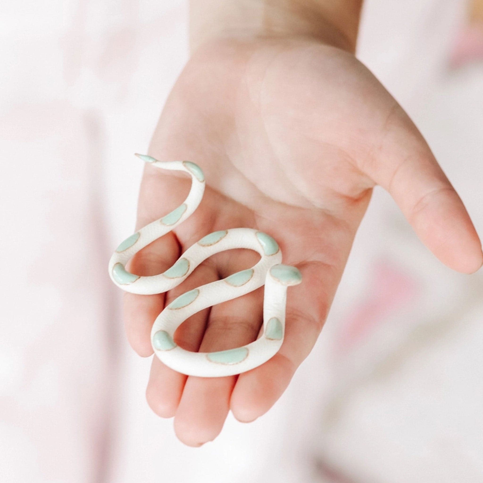 Sitting on the palm of a person’s hand is a white ceramic, cream snake. The snake&#39;s body twists multiple times from head to tail. There are brown bordered turquoise circles on the top of the snake. 