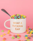 Classic white ceramic mug labeled 'What a Fucking Day' in gold shimmering capital lettering. Mug has a round white handle fasted on the left hand side. Mug is filled with overflowing, rainbow colored fruit loops. Behind lays a bright pink background.