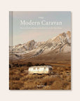 Hard cover of travel book titled 'The Modern Caravan, stories of love, beauty, and adventure on the open road' in white pressed lettering. Behind the title is a cool toned mountain scene. Below the mountains sits a silver airstream trailer amidst an open valley.