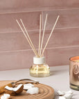 A oil diffuser with a clear glass filled with scented oil a clean cream colored lid and five neutral wood reeds stemming out from the top.