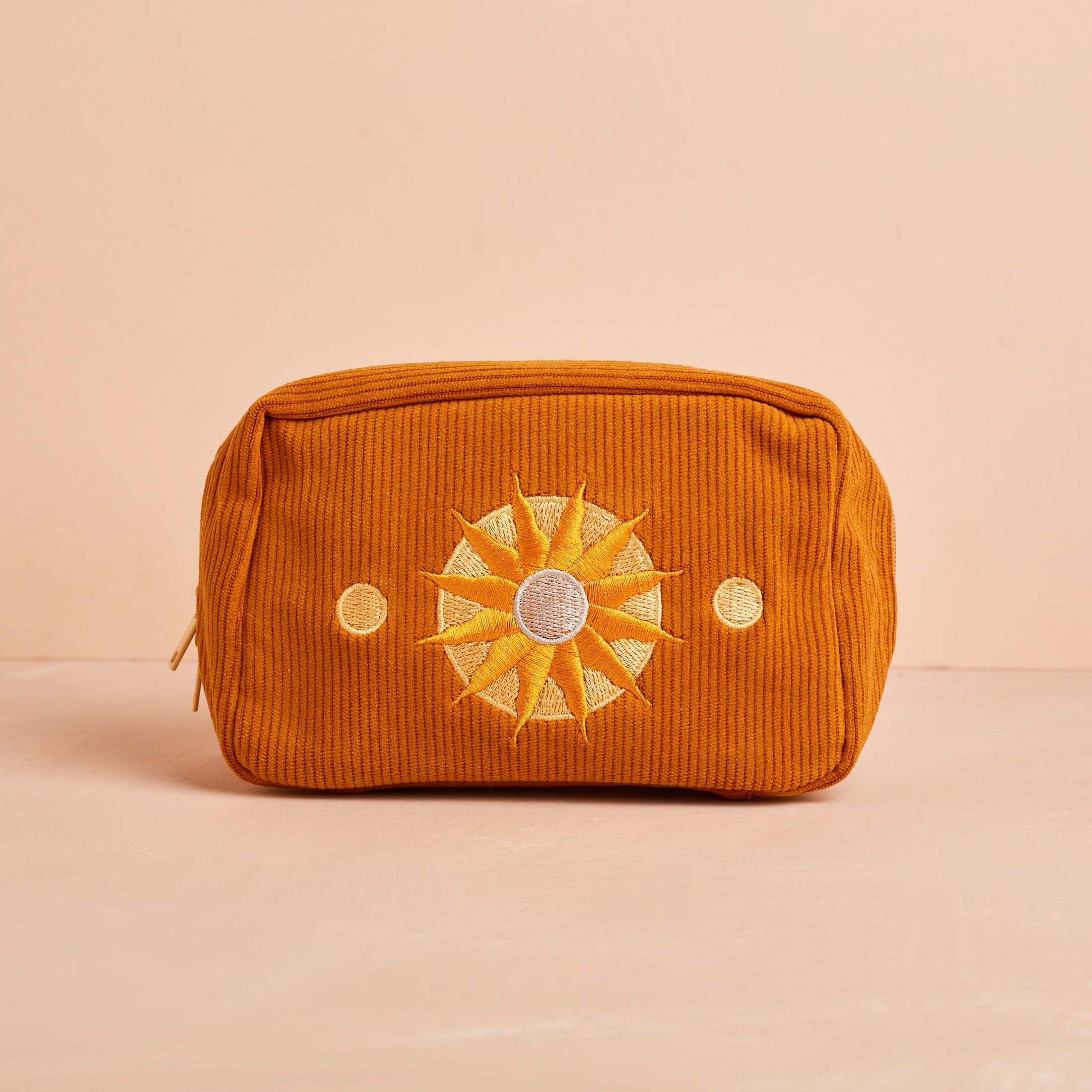 A burnt orange corduroy makeup bag with a sun design and two circles beside it.