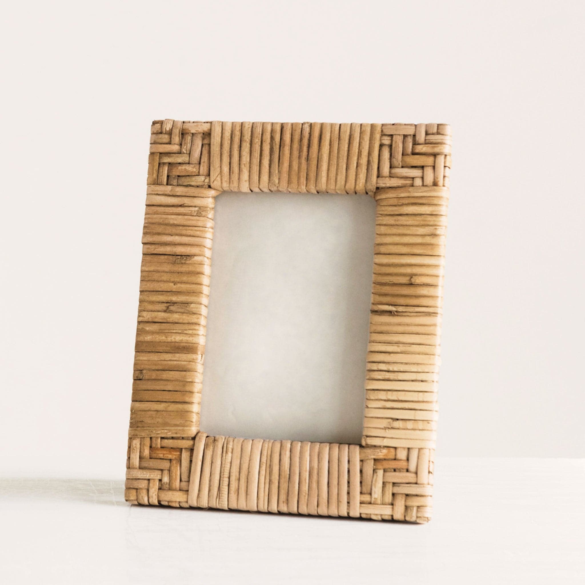 In front of a white background is a rattan picture frame. The corners of the picture frame are a chevron design and the sides are stripes. 