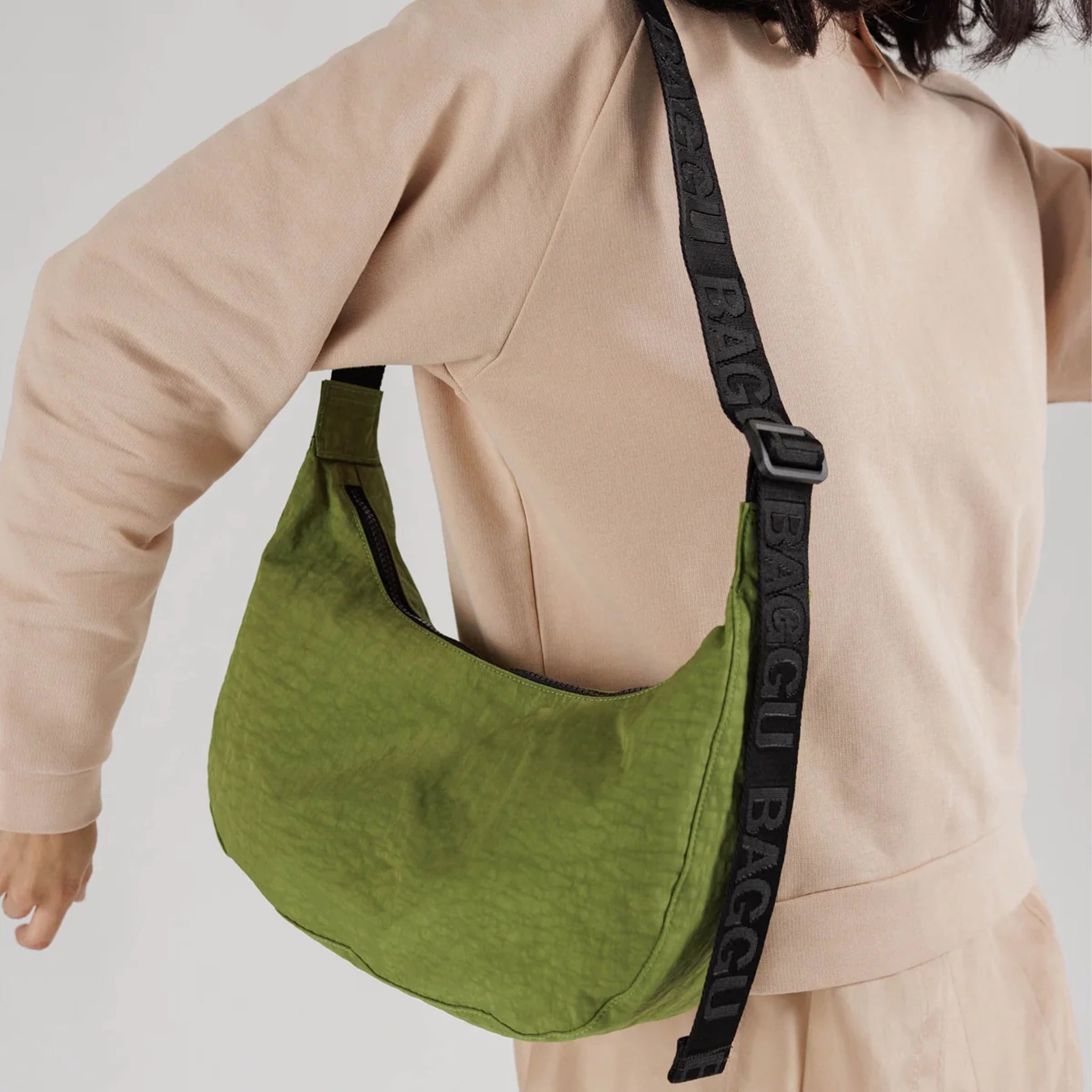 A vibrant green nylon crescent shaped bag with an adjustable black strap that reads, "BAGGU" repeated on the strap and a single zipper.