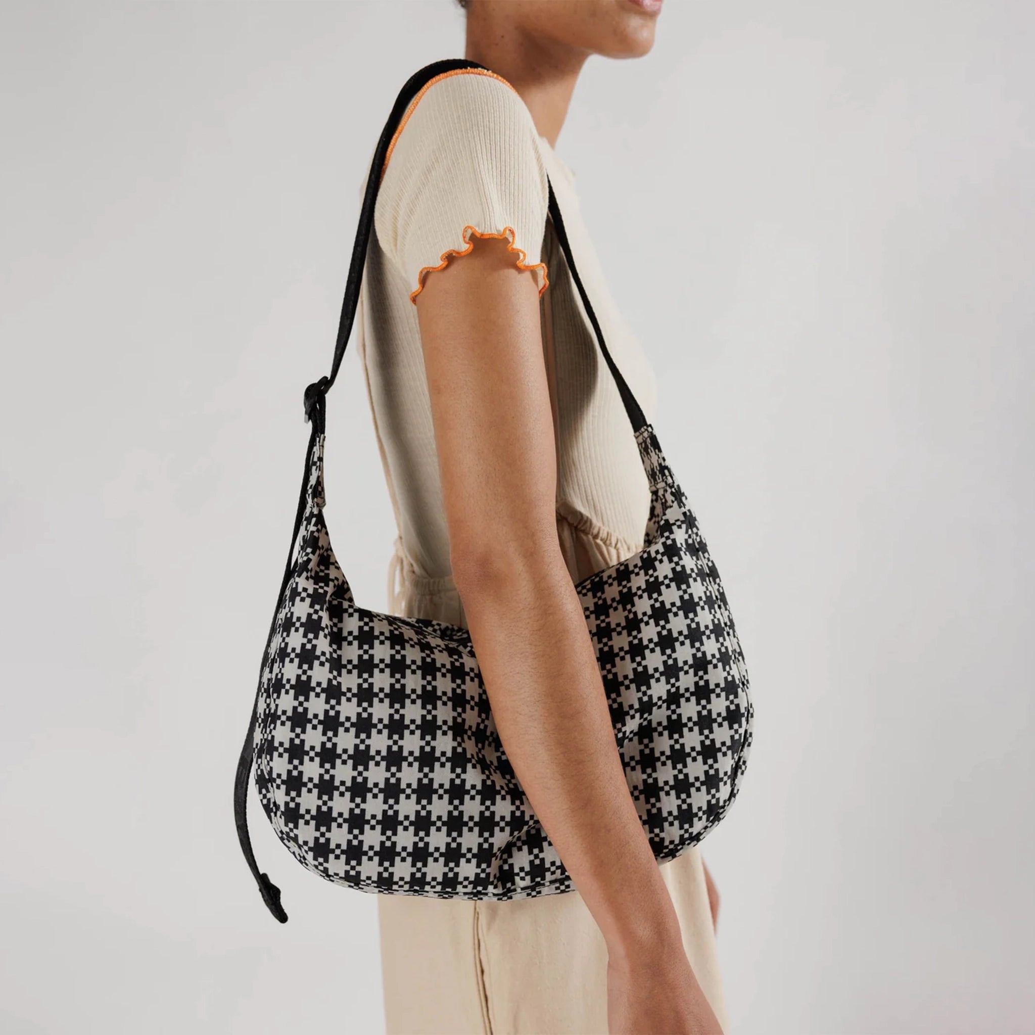 A black and white gingham nylon crescent shaped bag with an adjustable black strap that reads, "BAGGU" in black and a single zipper.