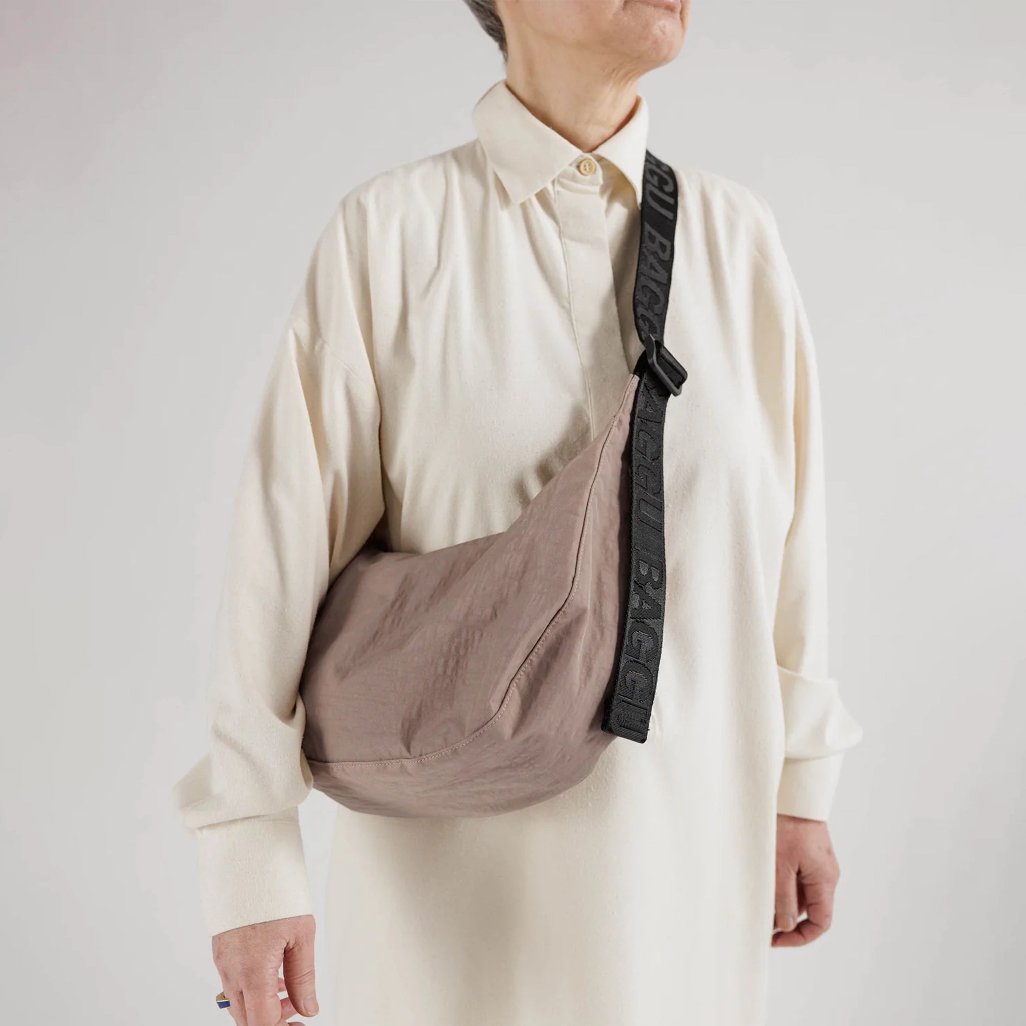 A taupe nylon crescent shaped bag with an adjustable black strap that reads, &quot;BAGGU&quot; in black and a single zipper.