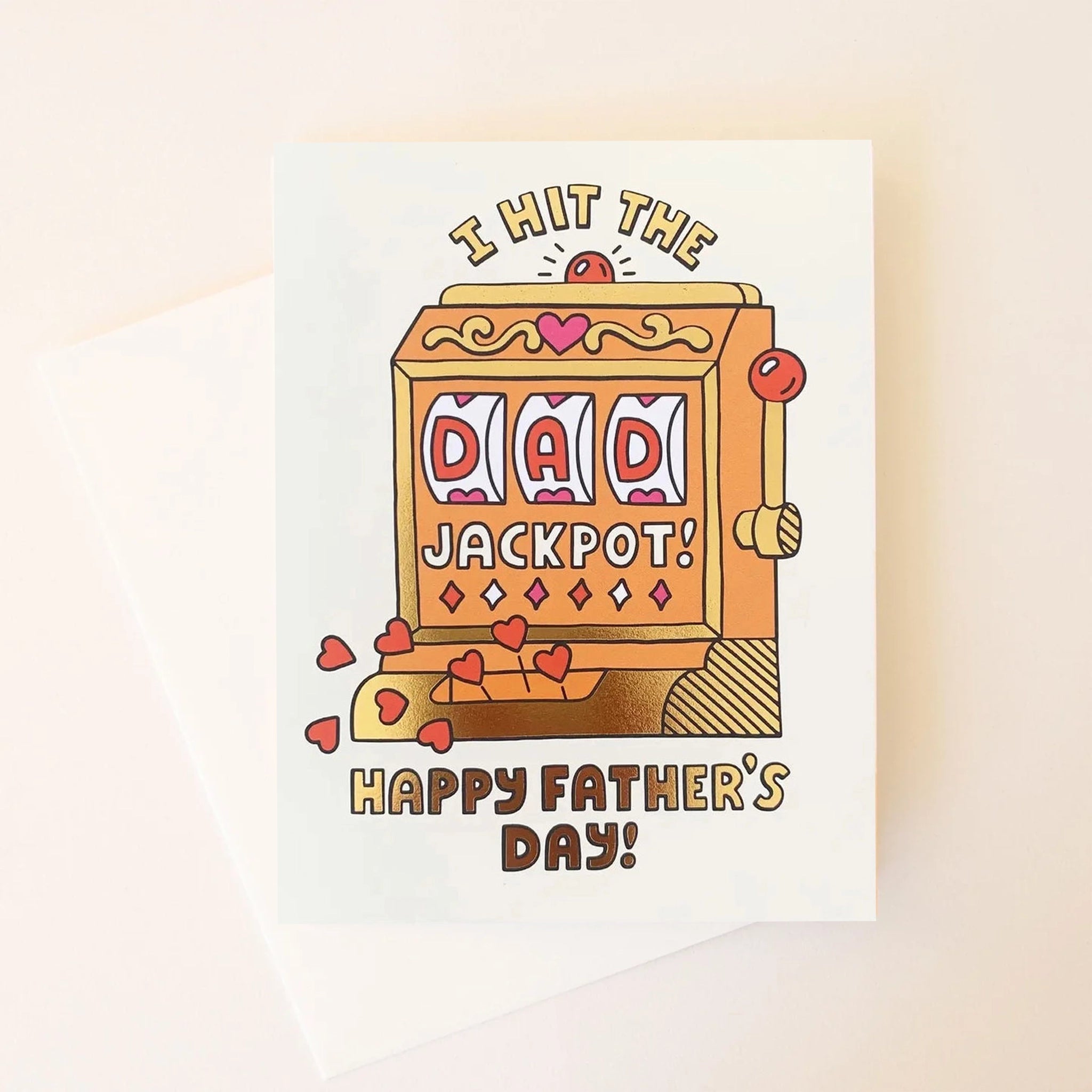 On a cream background is a white card with an orange and gold slot machine that&#39;s detailed with hearts and reads, &quot;I hit the DAD jackpot! Happy Father&#39;s Day!&quot;. &quot;DAD&quot; is written in the slot machine as the winning numbers. Also included is a coordinating white envelope.