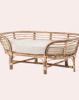 In front of a soft pink background is a rattan dog bed. The dog bed is an oval shape with six rattan legs on the bottom. Inside the bed is a light gray bed cushion.