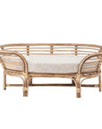 In front of a white background is a rattan dog bed. The dog bed is an oval shape with six rattan legs on the bottom. Inside the bed is a light gray bed cushion.