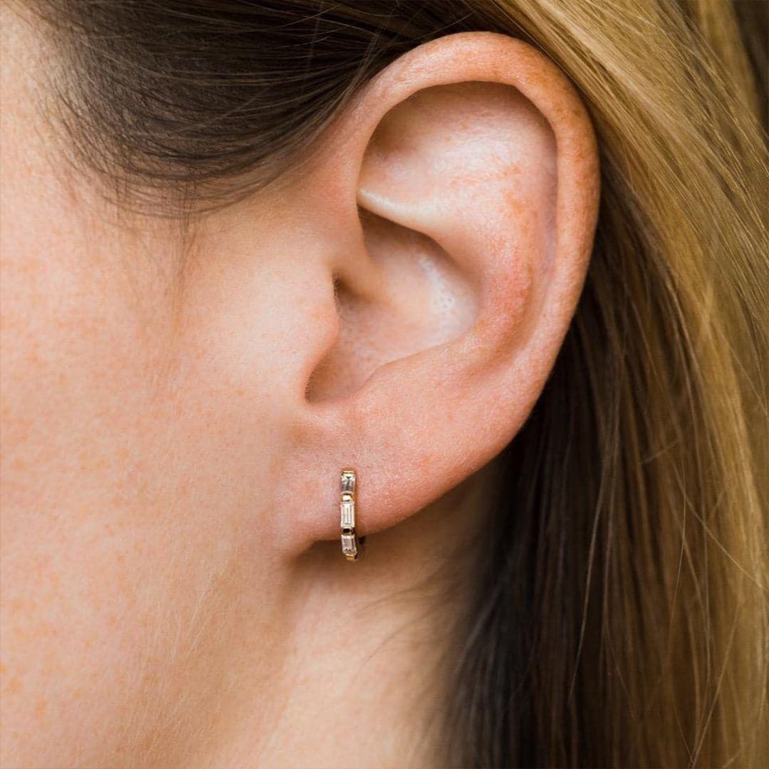 A small huggie hoop earring worn on a model's ear and features baguette style cubic zirconias along the gold huggie earring.