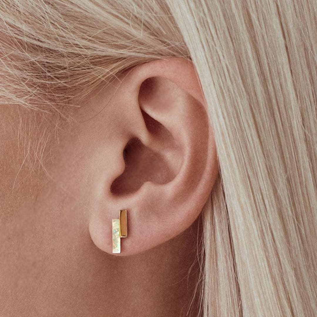 On a model's ear is a stud earring with two vertical bars staggered against one another. One is opal and the other is gold plated. 