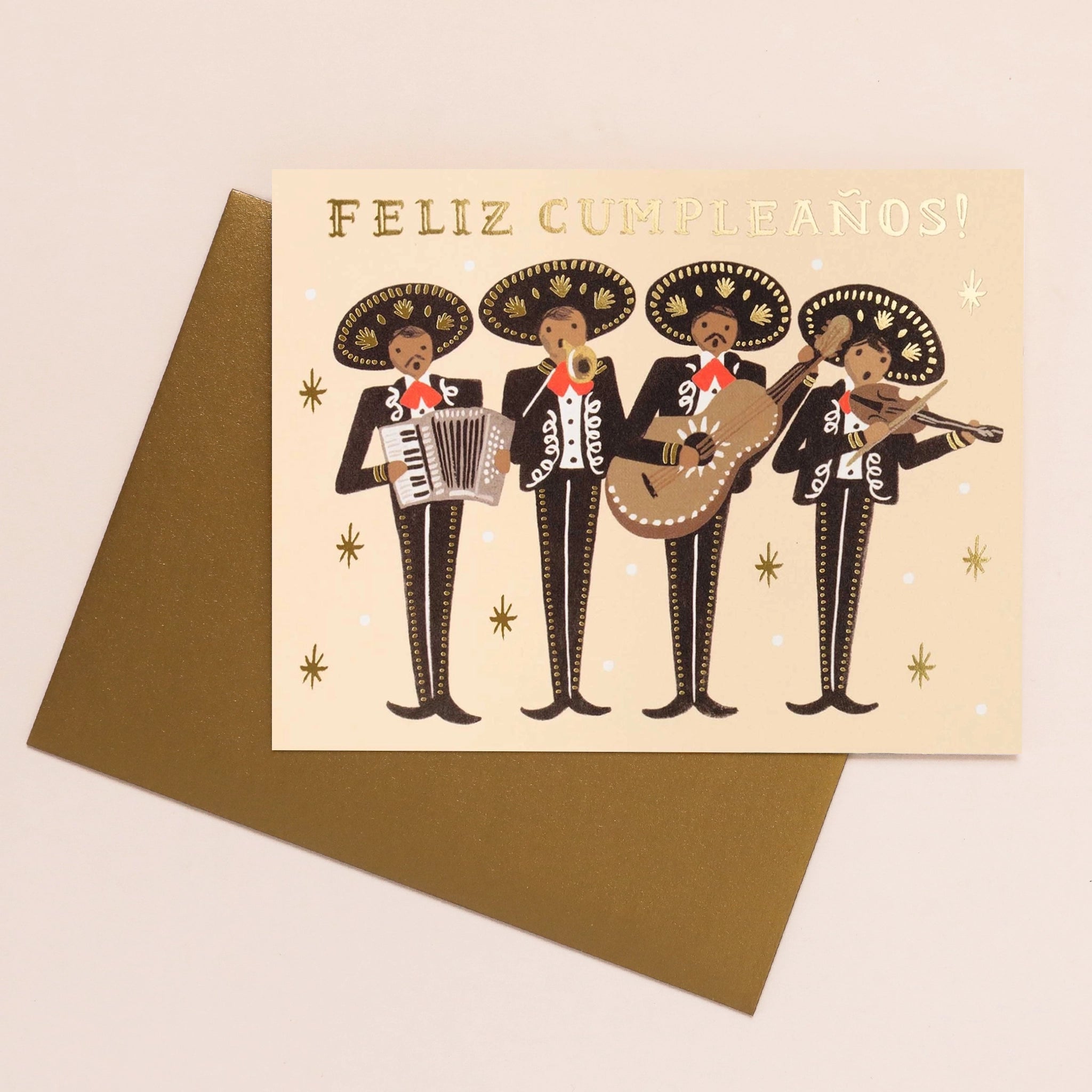On a cream background is a tan card with a four man mariachi band playing instruments with black suits hats with gold detailing along with gold foiled text at the top that reads, &quot;Feliz Cumpleaños!&quot;. It is also accompanies by a gold envelope.