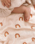 Neutral toned swaddle with warm rainbow design displayed under a baby with sweet button toes and a tan ruffly skirt.
