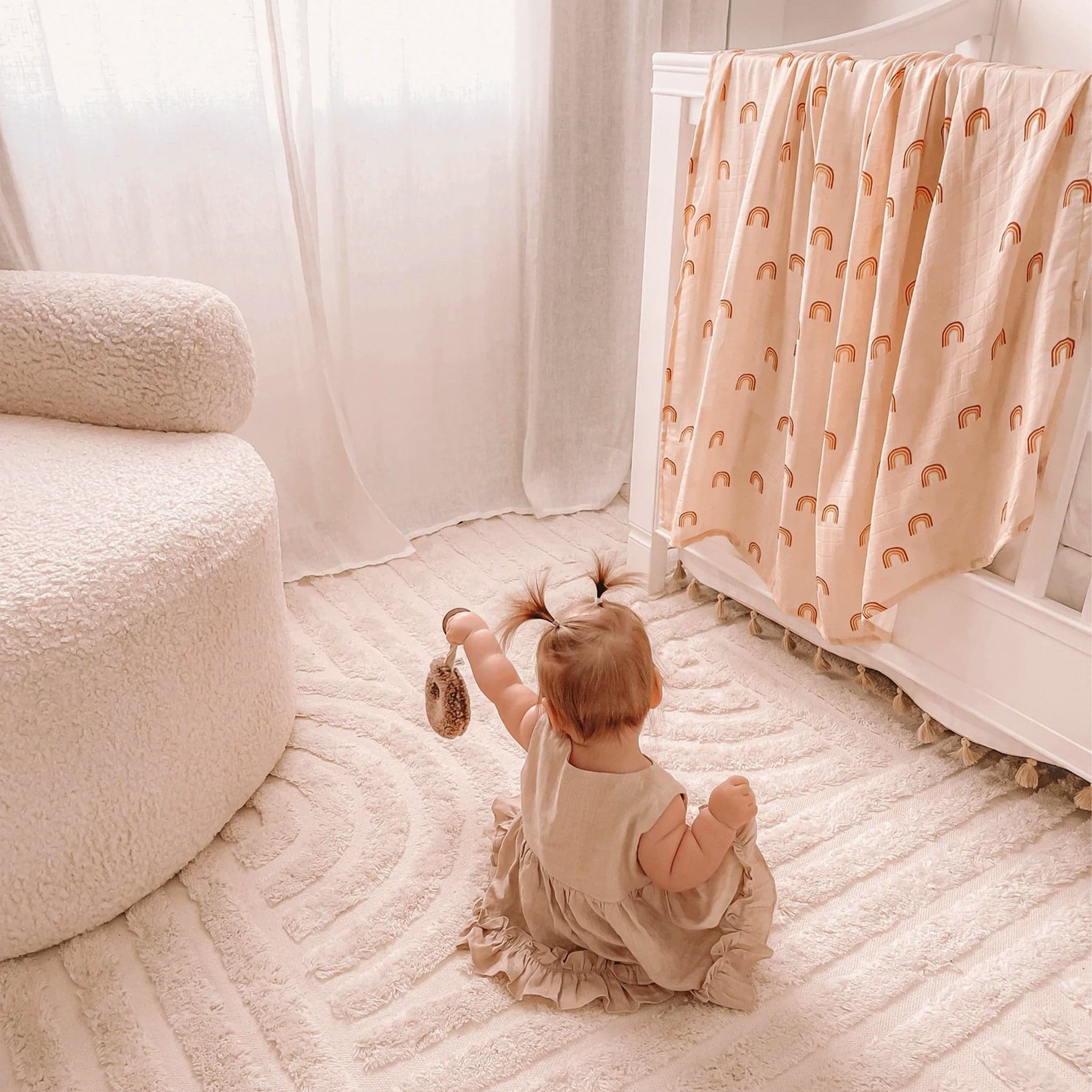Peach swaddle with warm toned rainbows displayed over crib edge. A small baby with two sprout pig tails sits besides the crib on a neutral toned rug. A white fuzzy chair sits to the left. 