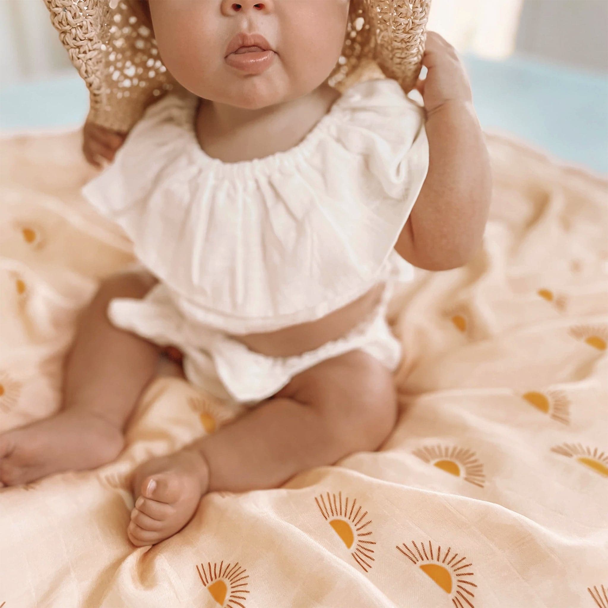 Pale beige swaddle with bohemian half sun pattern positioned as a changing cloth below a small baby. The baby is wearing a white ruffled set and woven sunhat. 