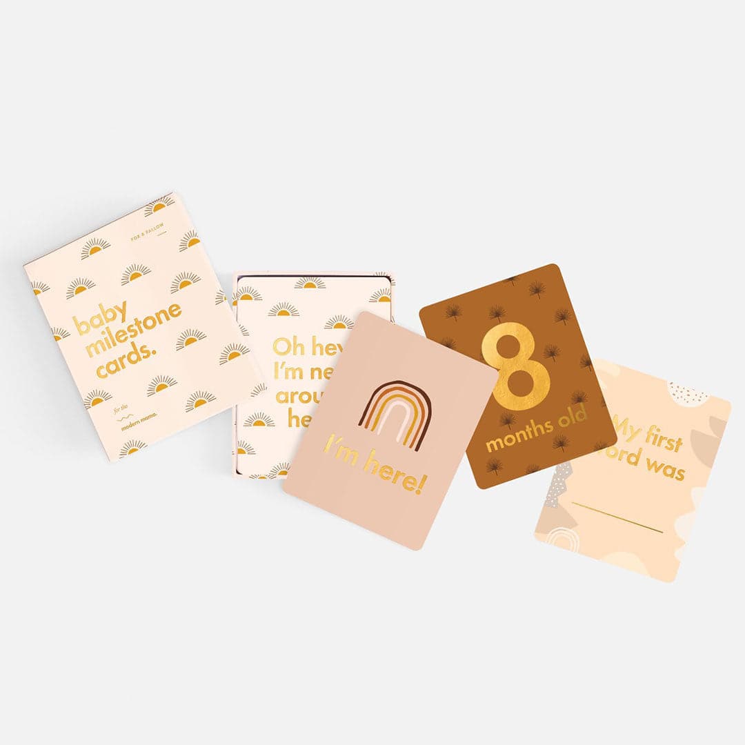 Ivory book with gold sun illustration and gold foil text &quot;Baby Milestone Cards - For the modern mama.&quot; With four cards pulled out, &quot;Oh hey I&#39;m new around here,&quot; &quot;I&#39;m here,&quot; &quot;8 months old,&quot; &quot;My first word was.&quot;