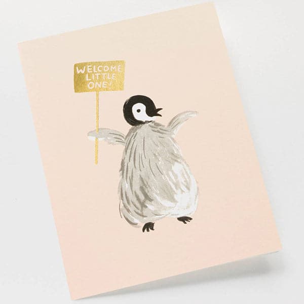 This adorable card is the color of a peach cloud. In the center is a grey feathery penguin chick holding a reflective gold sign labeled &#39;Welcome Little One&#39;. The card is accompanied by a solid white envelope.  Edit alt text