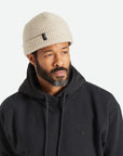 A knit ribbed beanie in a beige color with a folded brim and a black label on the front right left corner.