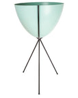 In front of a white background is a turquoise colored bullet planter. The planter has a wide top and narrows at the bottom. The planter is held up by a black metal stand. The stand has three black metal legs.