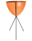 In front of a white background is an orange colored bullet planter. The planter has a wide top and narrows at the bottom. The planter is held up by a black metal stand. The stand has three black metal legs.