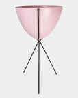 In front of a white background is a pink colored bullet planter. The planter has a wide top and narrows at the bottom. The planter is held up by a black metal stand. The stand has three black metal legs.