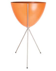 In front of a white background is an orange colored bullet planter. The planter has a wide top and narrows at the bottom. The planter is held up by a silver metal stand. The stand has three silver metal legs.
