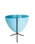 In front of white background is a bright blue planter in a short black metal stand. The bullet planter is wide at the top and narrow at the bottom. The metal stand has three legs.