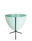 In front of white background is a turquoise planter in a short black metal stand. The bullet planter is wide at the top and narrow at the bottom. The metal stand has three legs.