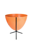 In front of white background is an orange planter in a short black metal stand. The bullet planter is wide at the top and narrow at the bottom. The metal stand has three legs.