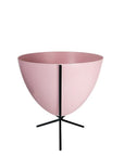 In front of white background is a soft pink planter in a short black metal stand. The bullet planter is wide at the top and narrow at the bottom. The metal stand has three legs.