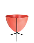In front of white background is a red planter in a short black metal stand. The bullet planter is wide at the top and narrow at the bottom. The metal stand has three legs.