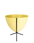 In front of white background is a yellow planter in a short black metal stand. The bullet planter is wide at the top and narrow at the bottom. The metal stand has three legs.