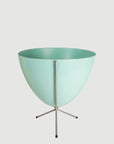 In front of white background is a turquoise planter in a short silver metal stand. The bullet planter is wide at the top and narrow at the bottom. The metal stand has three legs.