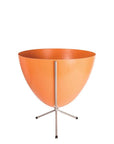 In front of white background is an orange colored planter in a short silver metal stand. The bullet planter is wide at the top and narrow at the bottom. The metal stand has three legs.