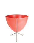 In front of white background is a red colored planter in a short silver metal stand. The bullet planter is wide at the top and narrow at the bottom. The metal stand has three legs.