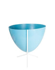 In front of white background is a bright blue bullet planter in a short white metal stand. The bullet planter is wide at the top and narrow at the bottom. The metal stand has three legs.