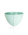 In front of white background is a turquoise bullet planter in a short white metal stand. The bullet planter is wide at the top and narrow at the bottom. The metal stand has three legs.