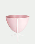 In front of white background is a pink bullet planter in a short white metal stand. The bullet planter is wide at the top and narrow at the bottom. The metal stand has three legs.