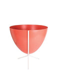 In front of white background is a red planter in a short white metal stand. The bullet planter is wide at the top and narrow at the bottom. The metal stand has three legs.