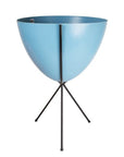 In front of white background is a bright blue planter in a black metal stand. The bullet planter is wide at the top and narrow at the bottom. The metal stand has three legs.