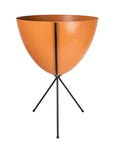 In front of white background is an orange planter in a black metal stand. The bullet planter is wide at the top and narrow at the bottom. The metal stand has three legs.