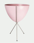 In front of white background is a soft pink planter in a silver metal stand. The bullet planter is wide at the top and narrow at the bottom. The metal stand has three legs.