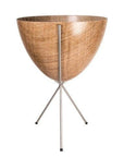In front of white background is a wood colored planter in a silver metal stand. The bullet planter is wide at the top and narrow at the bottom. The metal stand has three legs.