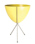In front of white background is a yellow planter in a silver metal stand. The bullet planter is wide at the top and narrow at the bottom. The metal stand has three legs.