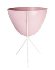 In front of white background is a soft pink planter in a white metal stand. The bullet planter is wide at the top and narrow at the bottom. The metal stand has three legs.