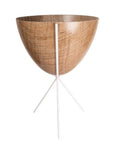 In front of white background is a wood colored planter in a white metal stand. The bullet planter is wide at the top and narrow at the bottom. The metal stand has three legs.
